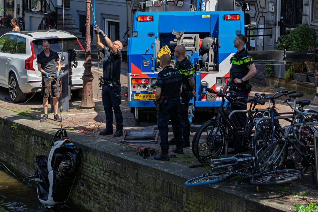 Police and crew recovering a vehicle from a canal in Amsterdam with a tow truck and bicycles nearby.