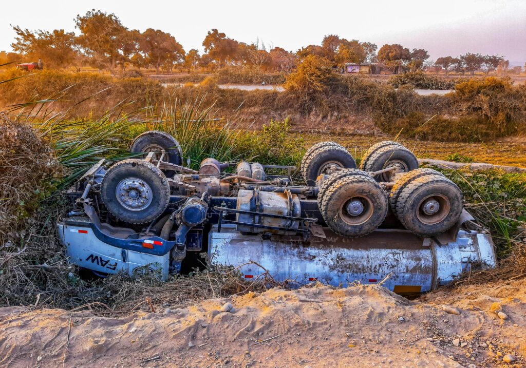 A truck overturned by the roadside on a rural dirt path during daytime.