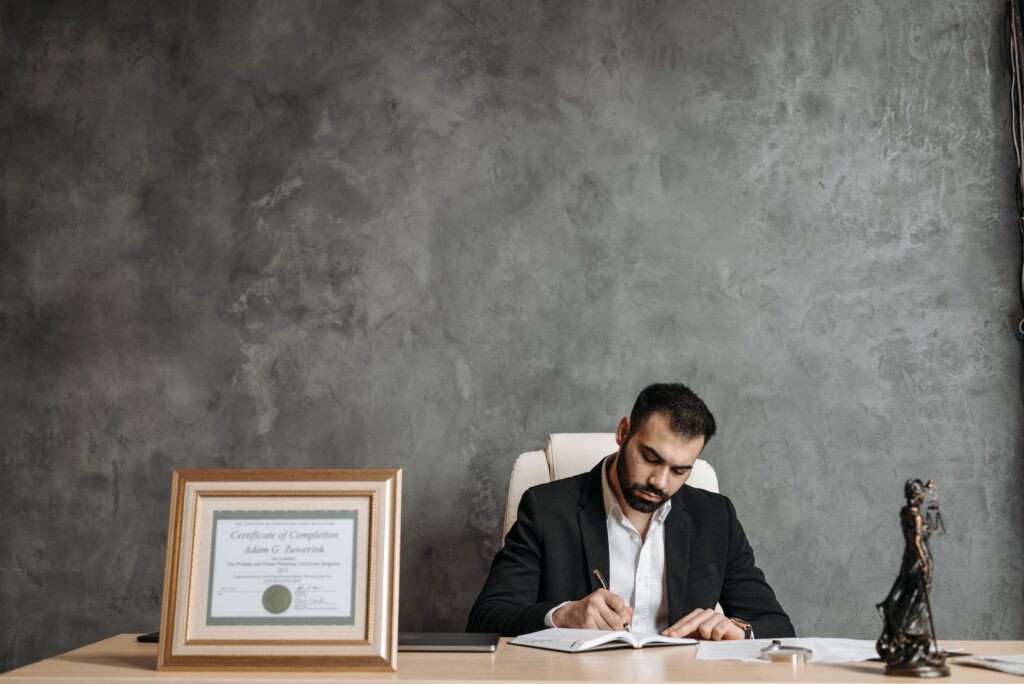 A business professional writing at a desk with a framed certificate and justice statue.