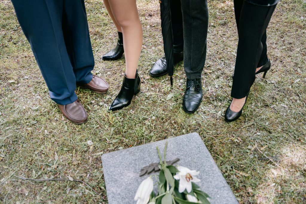 Shoes of mourners gathered around a gravestone with lilies in a cemetery.
