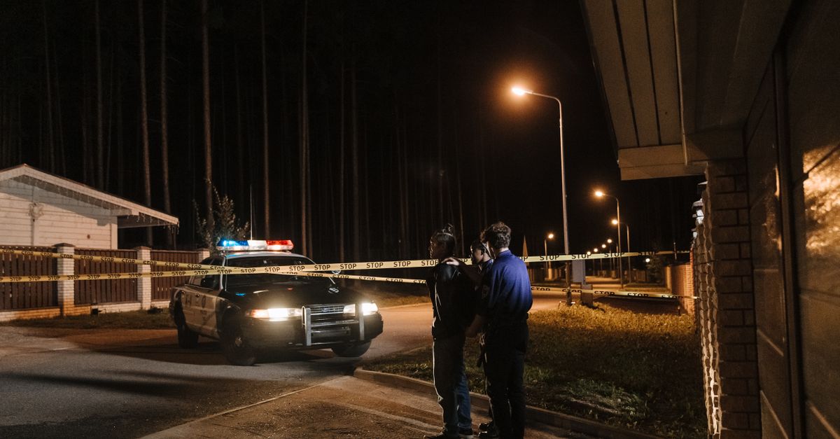 Police officers at a nighttime crime scene with a police car and caution tape under streetlights.