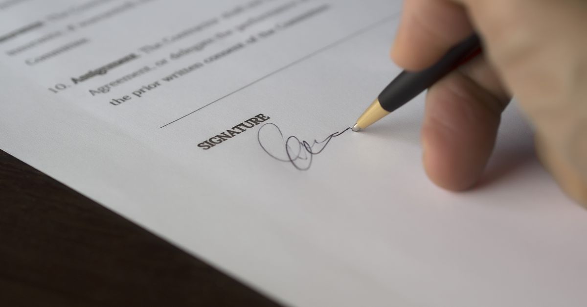 A hand signs a formal contract with a pen on a wooden desk.