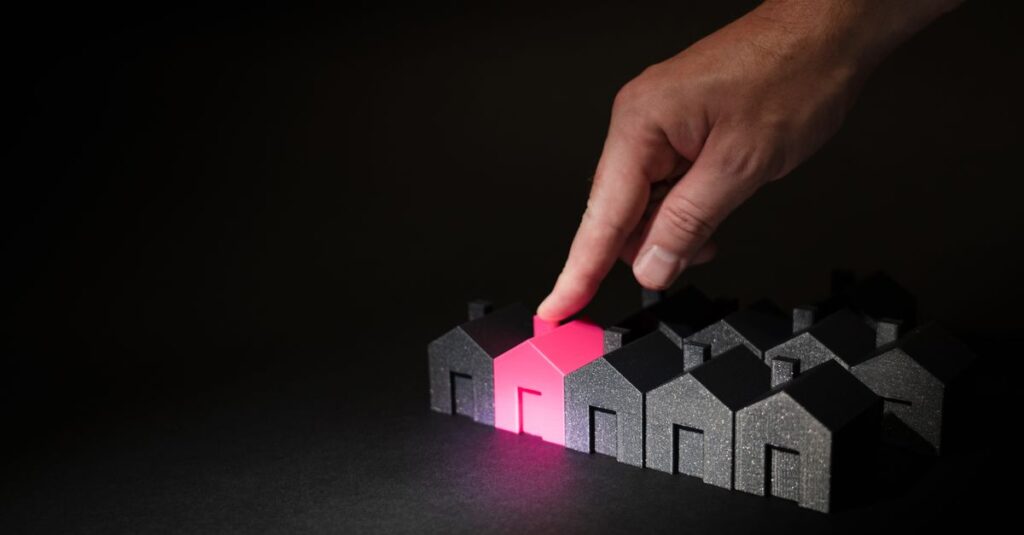 A hand selecting a glowing pink house among dark miniature houses, symbolizing real estate choices.