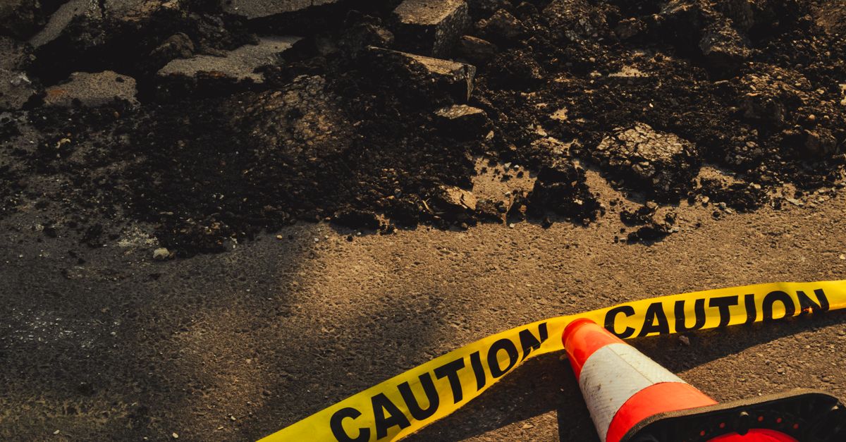 Broken sidewalk with rubble, caution tape, and traffic cone in urban construction site.