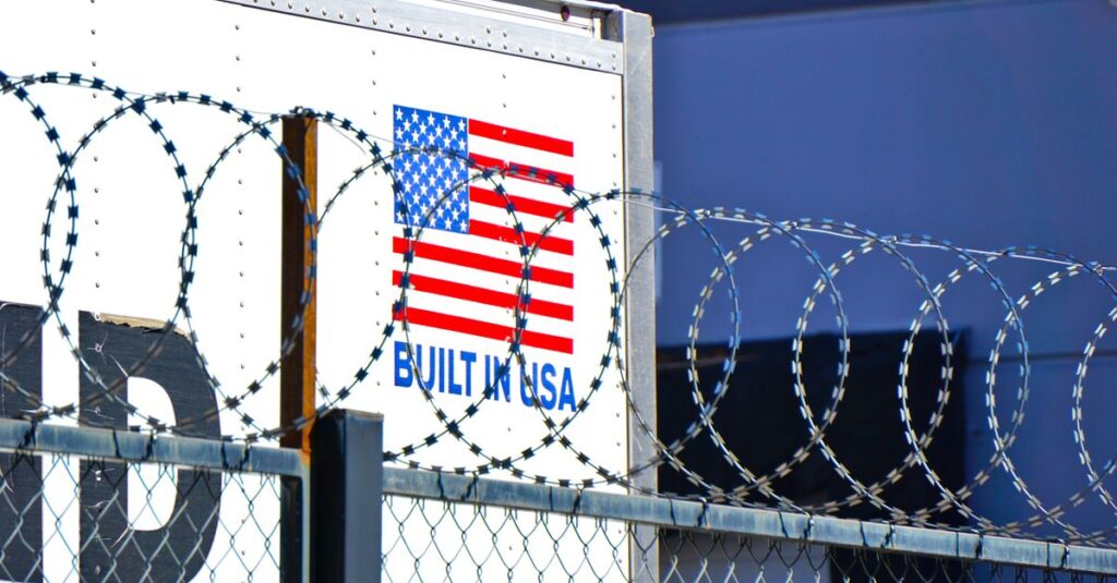 Industrial scene with barbed wire fence and American flag logo on building.