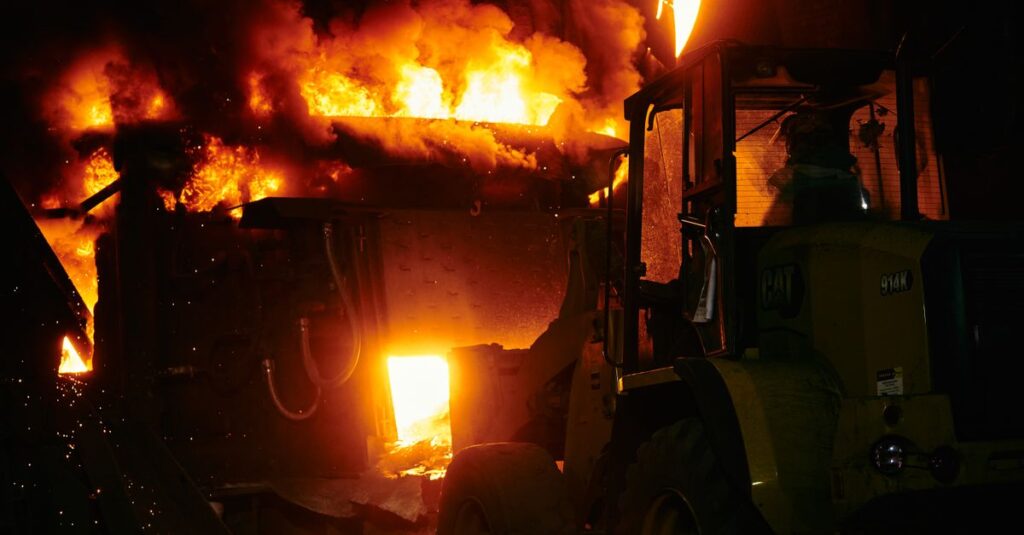 A powerful image of an intense industrial fire with a vehicle nearby, taken at night.