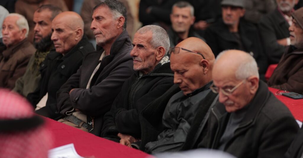 A group of senior men gathered at an outdoor community meeting during the day.