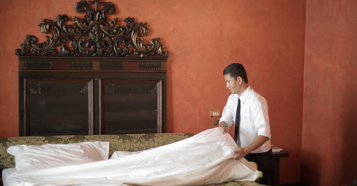 A hotel staff member in formal attire making a bed in a luxurious bedroom setting.