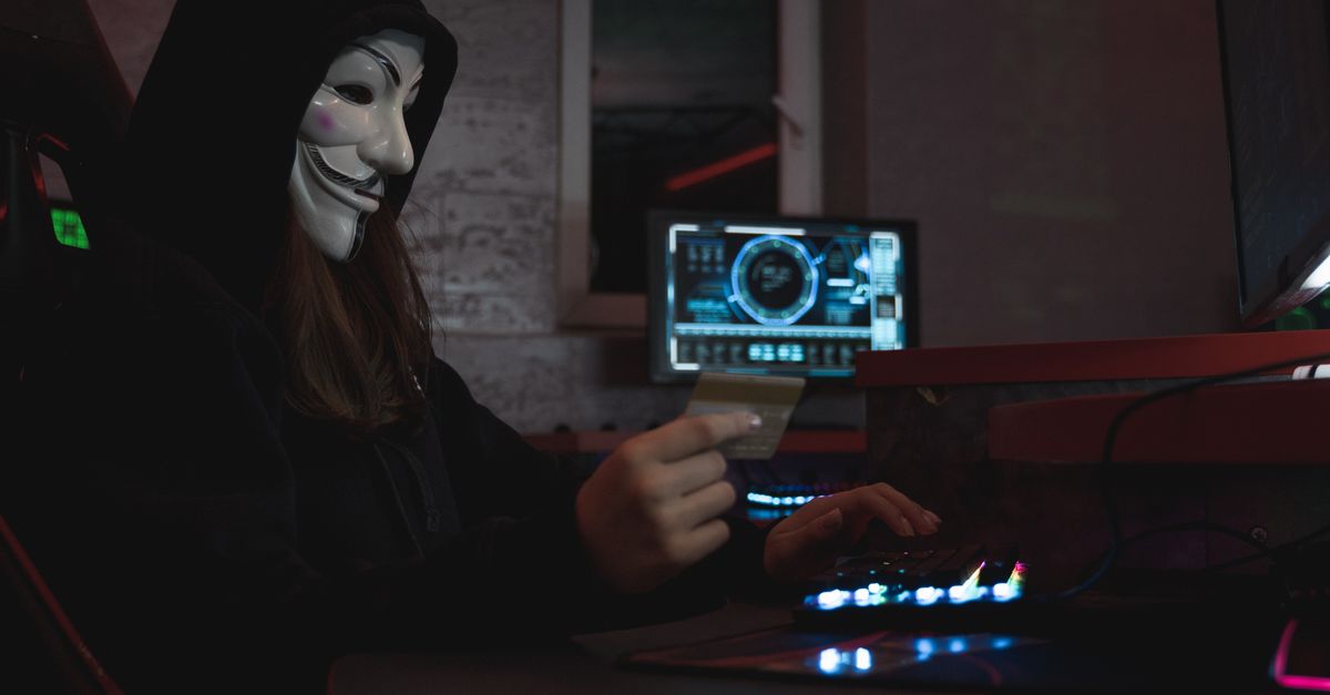 Masked hacker with credit card at computer, symbolizing cybercrime and anonymity.