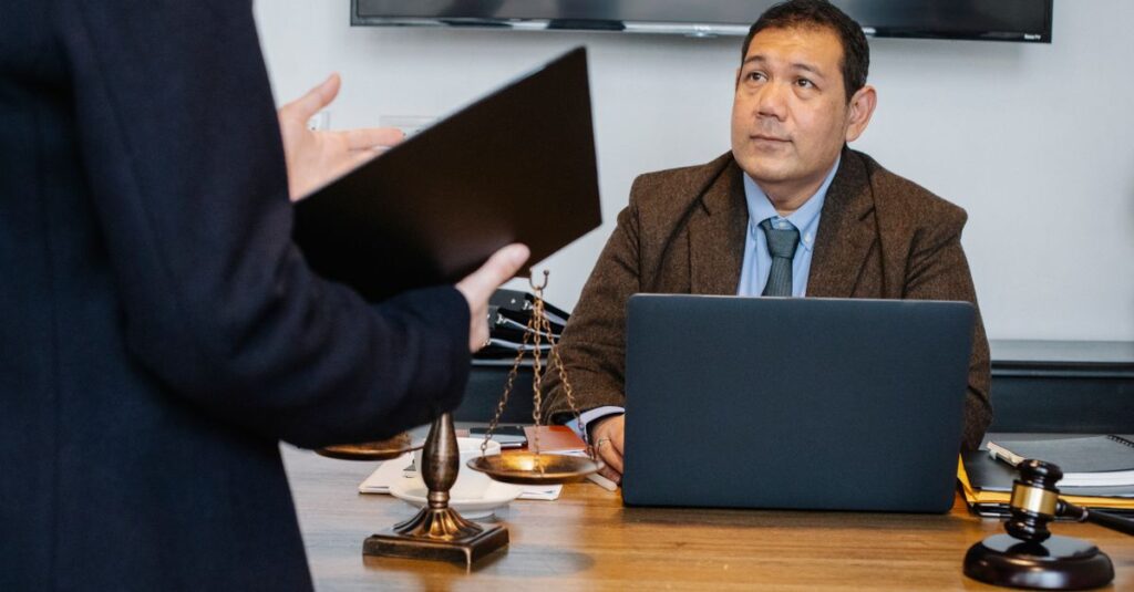 Crop unrecognizable employee representing new case details to concentrated middle aged ethnic lawyer sitting at table with laptop gavel and justice scales