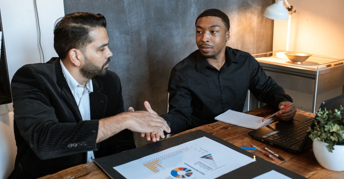 Two businessmen shaking hands over a desk with charts in a modern office.