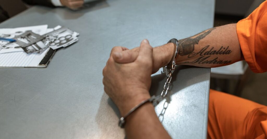 A handcuffed prisoner in an orange jumpsuit sits at a table during an interrogation.