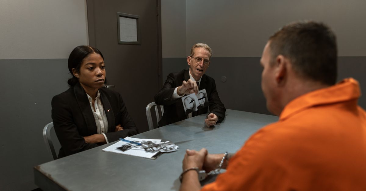 A suspect in an orange jumpsuit is interrogated by detectives in a dimly lit room.