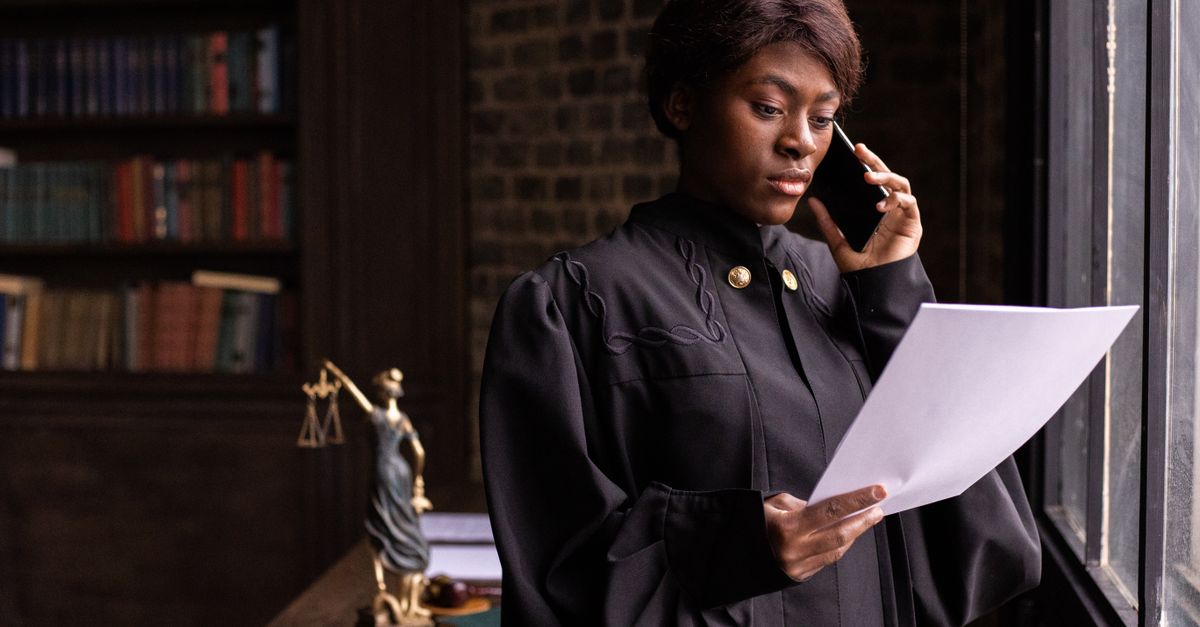 African American judge in robe, reading document and talking on phone by the office window.