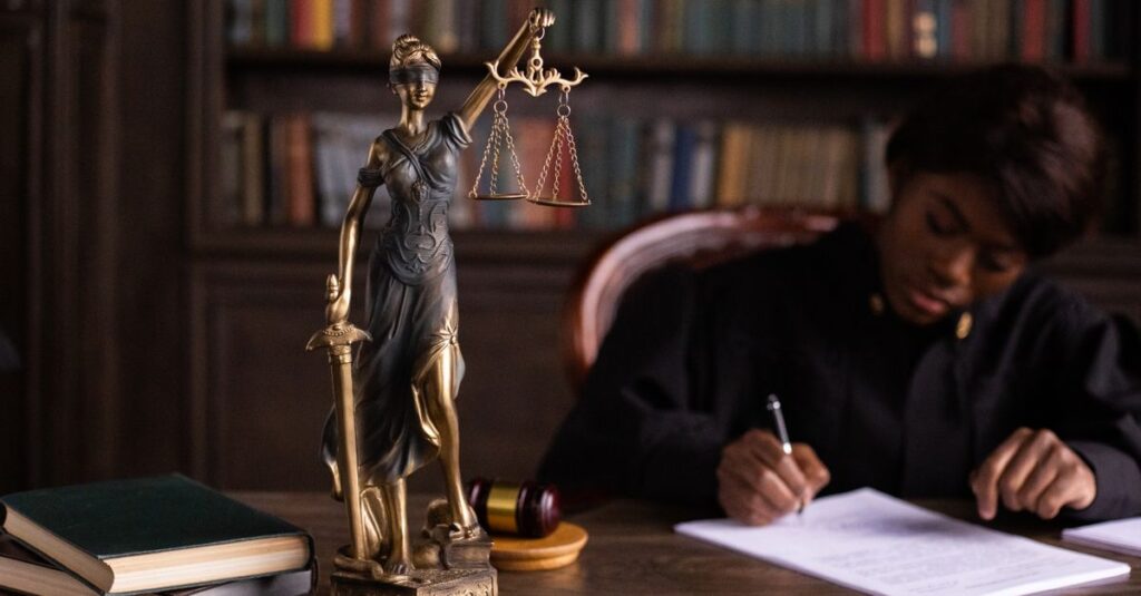 A focused judge writing on documents beside a Lady Justice statue in an office.