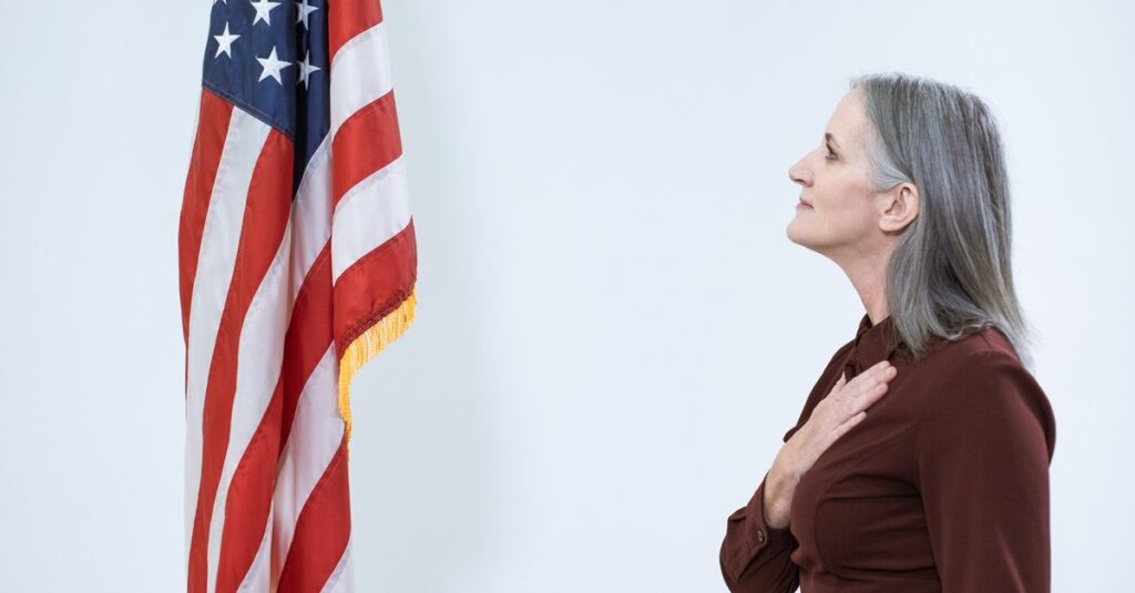 A senior woman pledges allegiance at a podium next to an American flag.