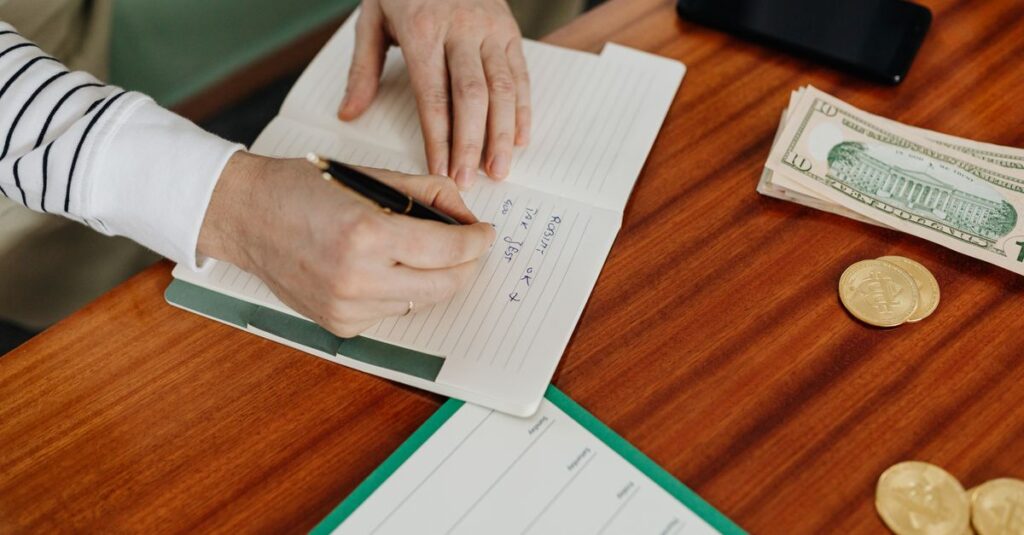Person writing in a notebook on a wooden table with cash, coins, and a phone nearby.