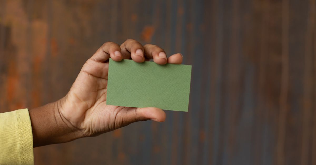 Unrecognizable African American person showing green mock up business card in hand while standing near wooden wall in modern studio
