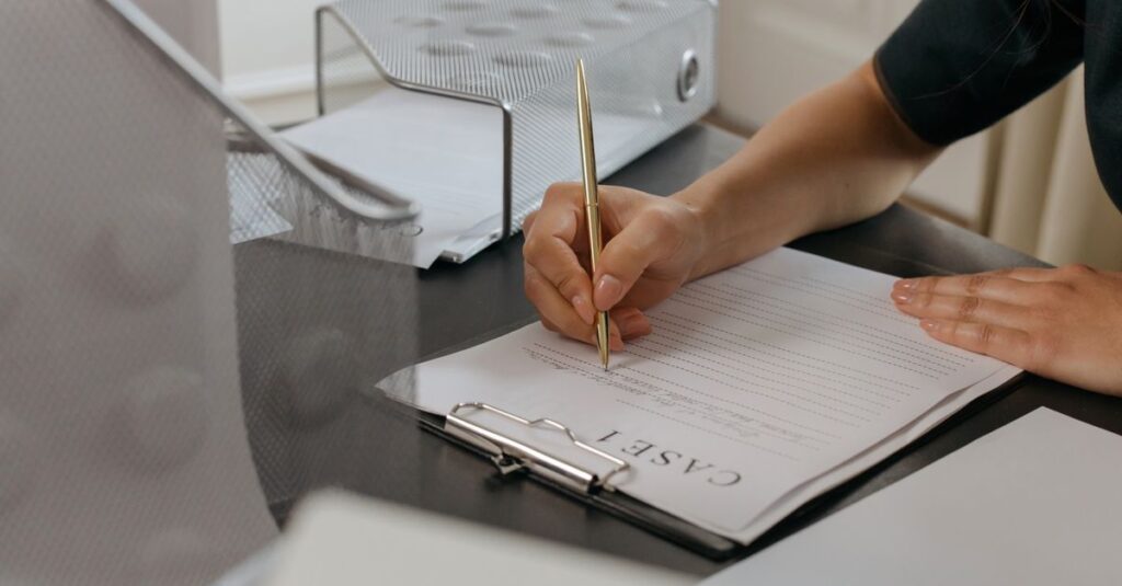 Close-up of a woman writing on documents with a gold pen in an office environment.