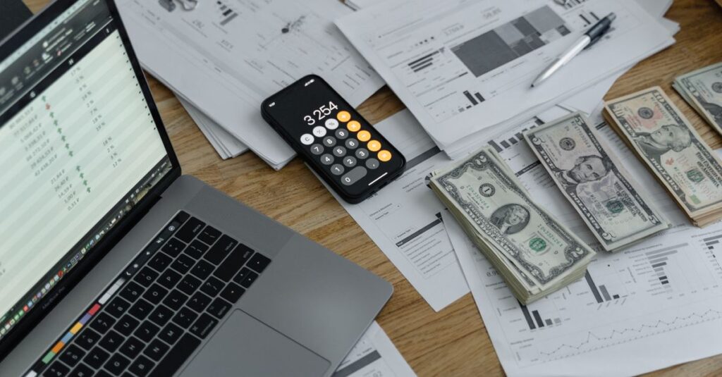 A desk with financial documents, currency, a laptop, and phone calculator.