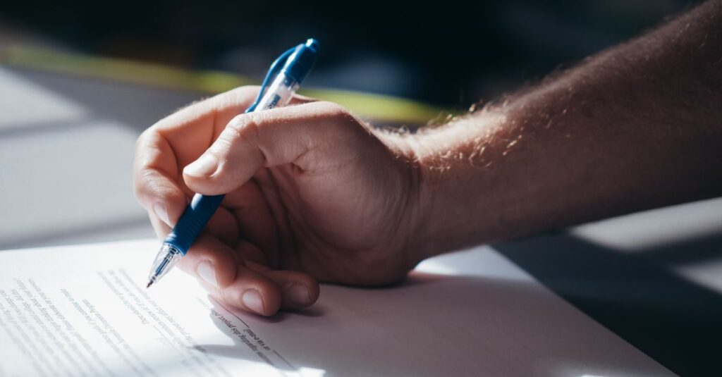 Detailed shot of a hand holding a blue pen while signing a document. Ideal for legal and business themes.