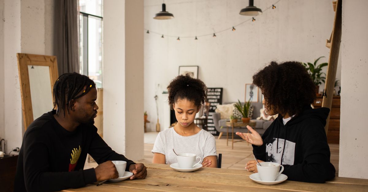 African American mother scolding unhappy daughter sitting at table with cups of coffee near father