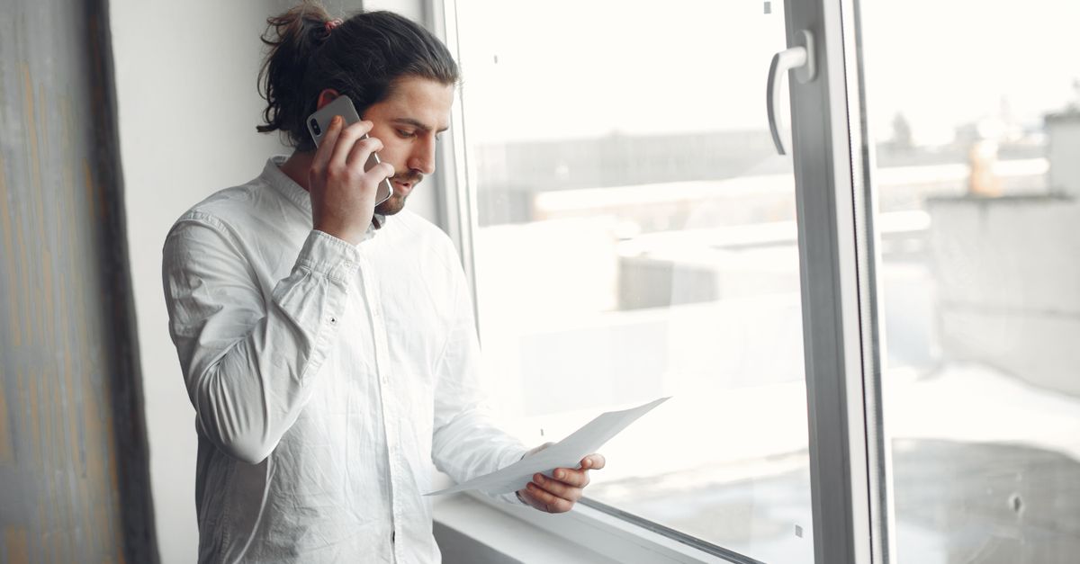 Young man in white shirt, on phone call holding a document, standing by a large window.