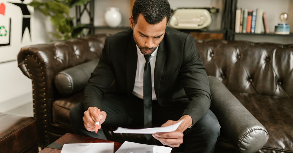 A businessman in a suit attentively reviews paperwork on a brown leather sofa.