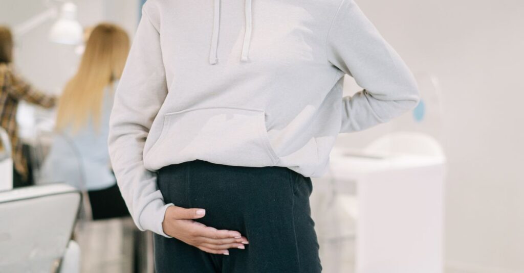 Pregnant woman standing in an office, hand on belly, wearing a hoodie. Modern workspace environment.