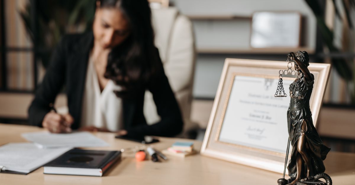 Focused woman writing at desk with Lady Justice figurine and certificate in office.