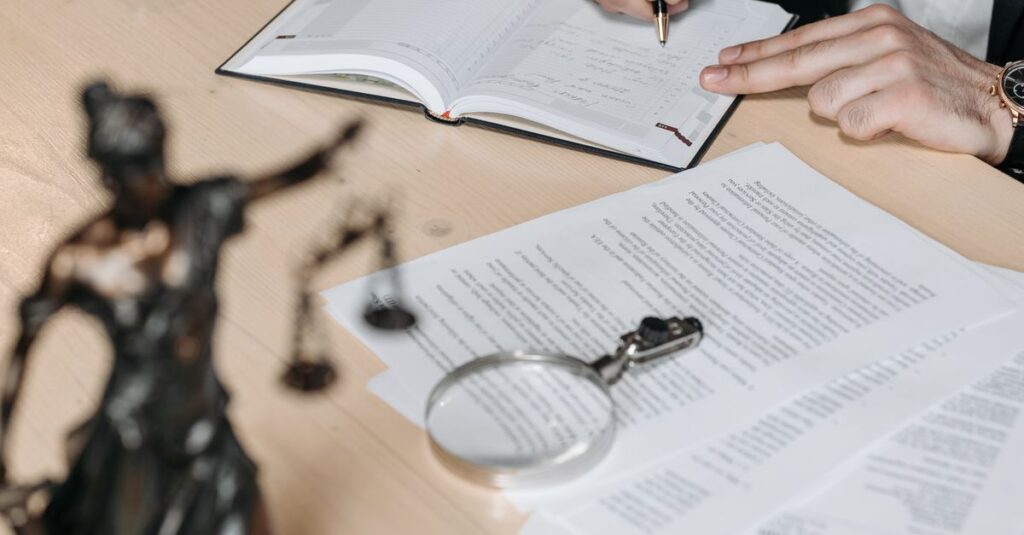 Professional working on documents at desk with legal statue and magnifying glass.