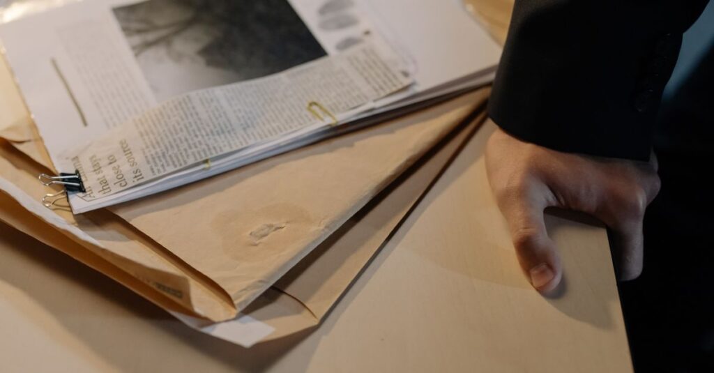Close-up of a hand resting on a desk near a stack of investigative documents and newspaper clippings.