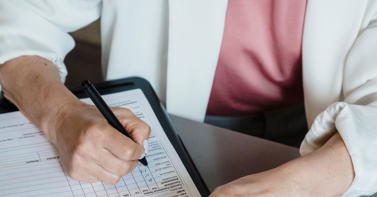 A professional woman in white jacket writing on a document clipboard during consultation.