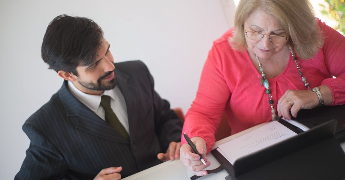 A senior woman signing paperwork with a young professional assisting her at a desk.