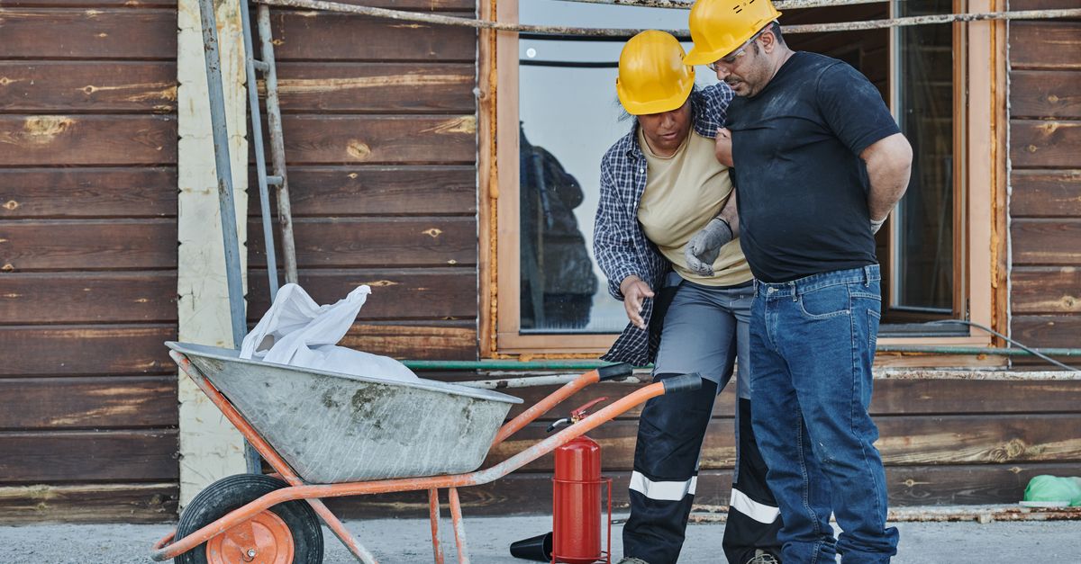 Two construction workers with hard hats assist an injured team member, highlighting workplace safety.