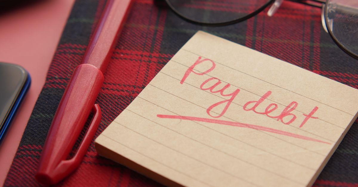 Close-up of a note reading 'Pay debt' next to a red pen on a plaid fabric, emphasizing financial reminders.