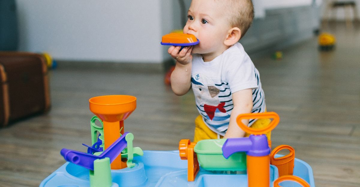 Adorable toddler chewing on toy piece while playing indoors with bright colored plastic toy.