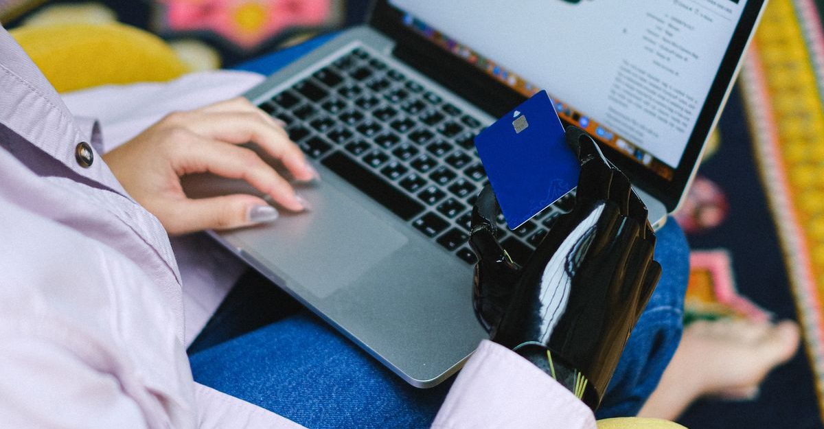 A person using a prosthetic arm shops online with a laptop and bank card indoors.