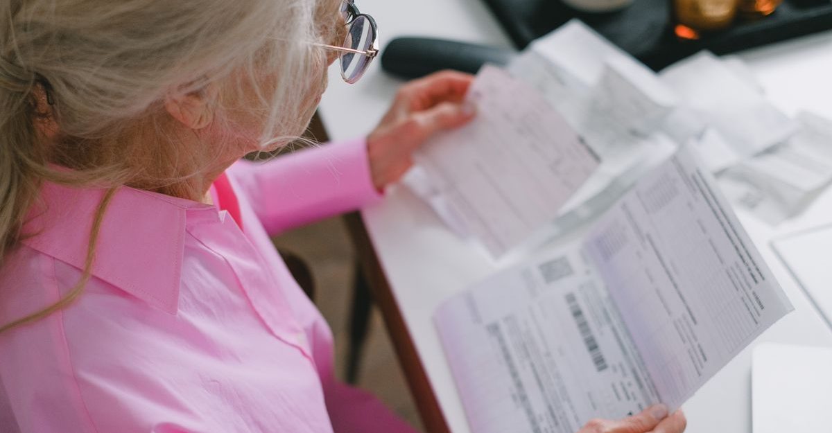 An elderly woman in glasses holds and reads important papers at a table indoors.