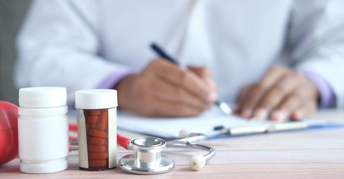 Close-up of doctor writing prescription with medicines and stethoscope on desk.